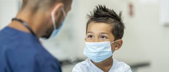 A child wearing a facemask looks up at a doctor during a visit.