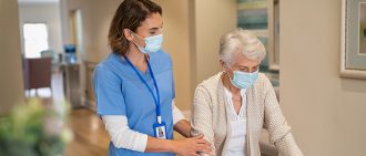 A smiling nurse with a face mask helps an older woman to walk around the nursing home with a walker.